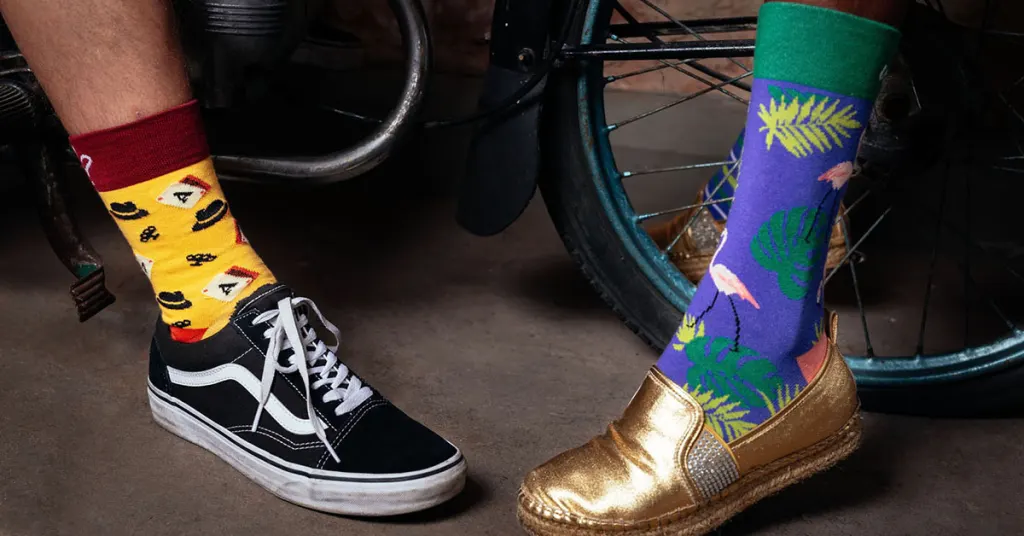 Dwie nogi przy kole roweru: jedna w skarpetce La Cosa Nostra i czarnych trampkach Vans, druga w skarpetce Flamingo i złotych espadrylach